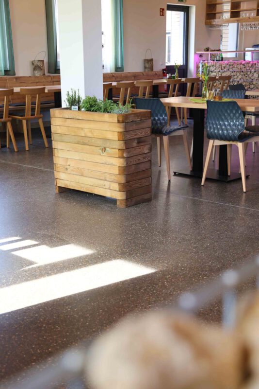 Restaurantbereich mit Holztischen und Stühlen, einem Pflanzkasten aus Holz in der Mitte und einem glänzenden Terrazzo-Boden, der Sonnenstrahlen reflektiert.