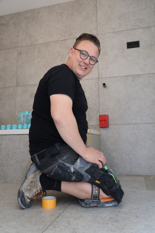 Fliesenleger Tim Dezius kniet lächelnd auf dem Boden vor großformatigen Wandfliesen, trägt Arbeitskleidung und Knieschoner. Arbeitsmaterialien sind im Hintergrund sichtbar.