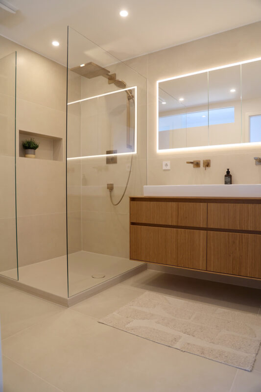 Modernes Badezimmer mit einer offenen, bodengleichen Dusche, beigen Wand- und Bodenfliesen, hölzernem Waschtischunterschrank, beleuchtetem Spiegel und minimalistischem Design.