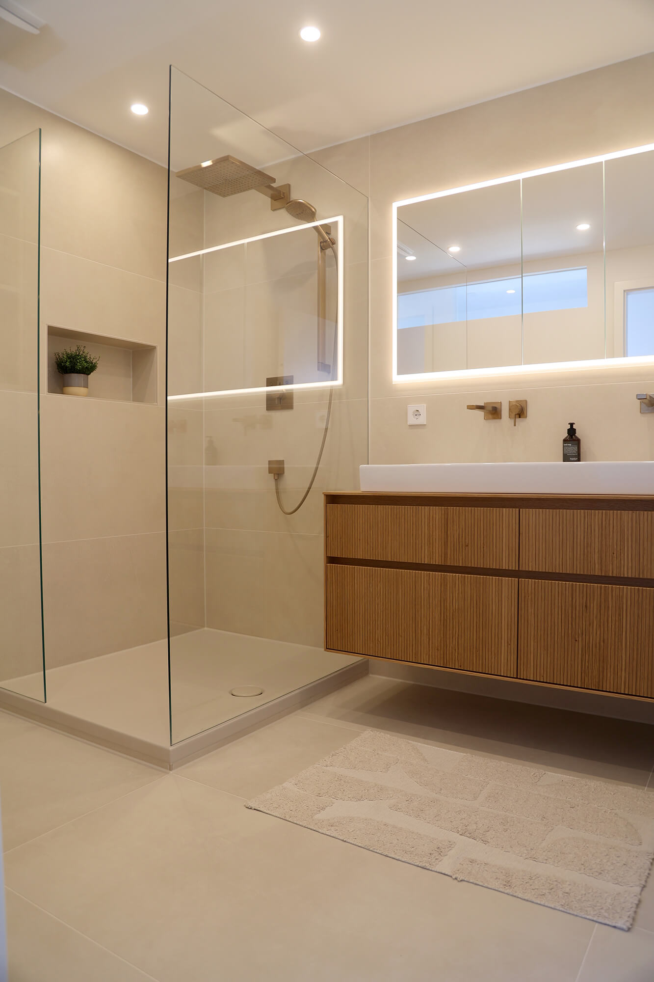 Modernes Badezimmer mit einer offenen, bodengleichen Dusche, beigen Wand- und Bodenfliesen, hölzernem Waschtischunterschrank, beleuchtetem Spiegel und minimalistischem Design.