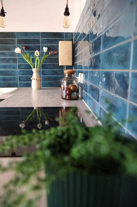 Moderne Küchenzeile mit glänzenden blauen Wandfliesen, einer Vase mit Tulpen, Glasbehälter für Kaffeekapseln und hängenden Vintage-Glühbirnen. Im Vordergrund eine grüne Zimmerpflanze.