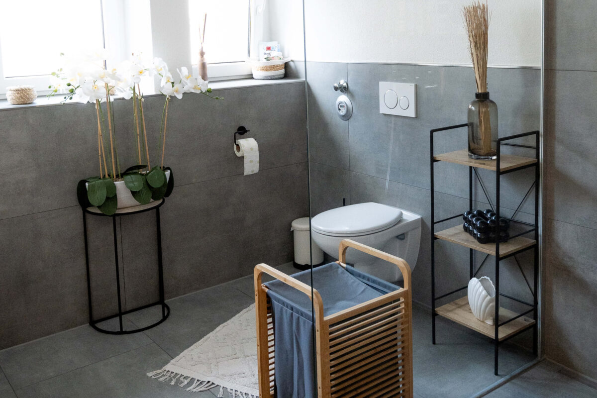 Modernes Badezimmer mit grauen Wand- und Bodenfliesen, dekoriert mit Orchideen auf einem schwarzen Metalltisch, einem Holzregal mit Dekoobjekten und einem Wäschekorb aus Holz.