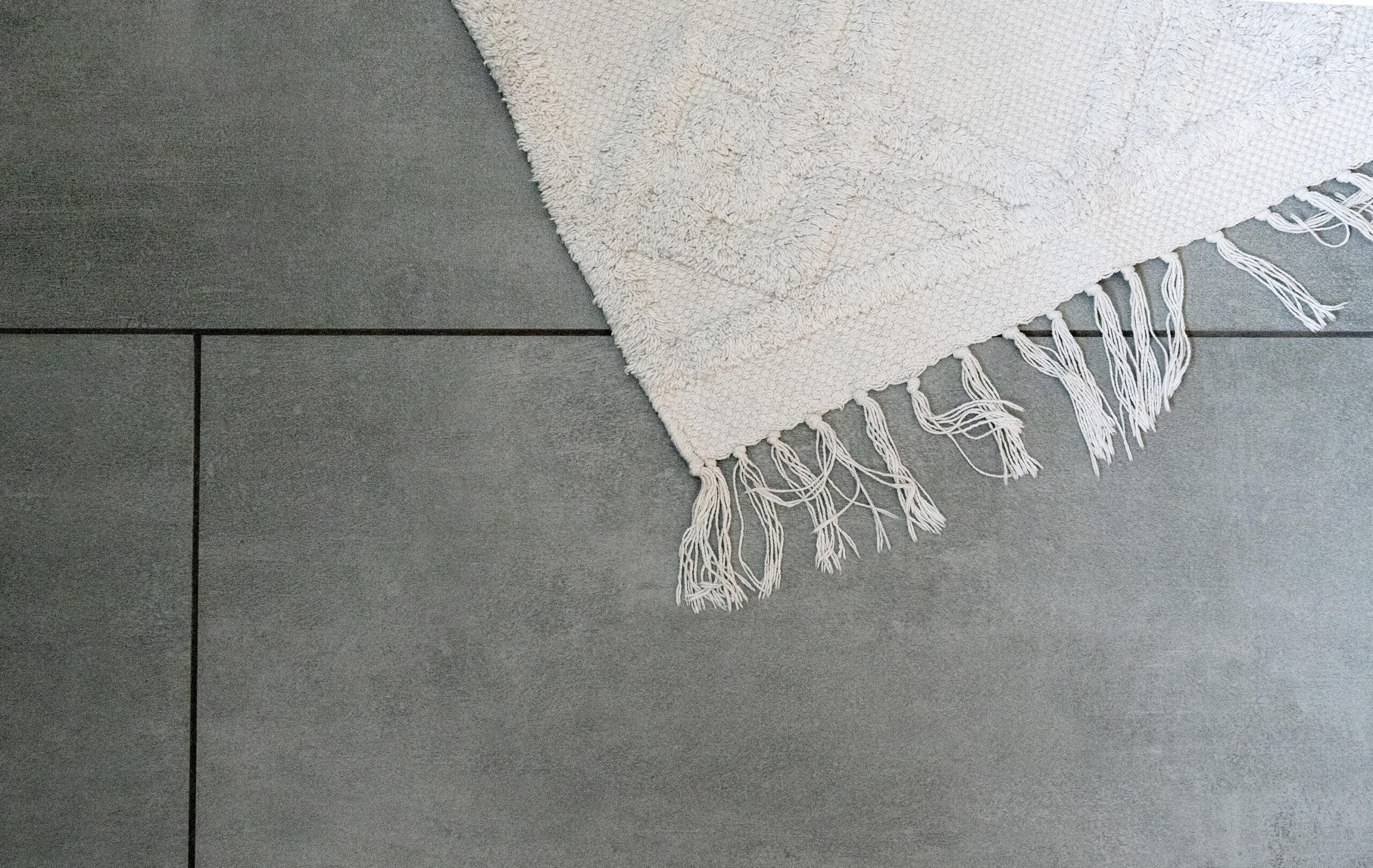 Detailaufnahme von grauen Fliesen mit feiner Struktur, kombiniert mit einem weißen, gefransten Teppich.