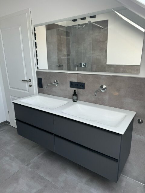 Modernes Badezimmer mit Doppelwaschbecken in einem dunklen Unterschrank, großem Spiegel und grauen Wand- und Bodenfliesen.