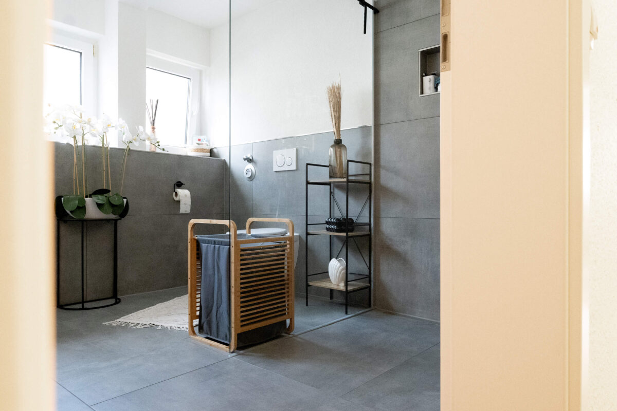 Modernes Badezimmer mit grauen, großformatigen Fliesen, einer offenen Dusche, dekorativen Pflanzen und einem Wäschekorb aus Holz.
