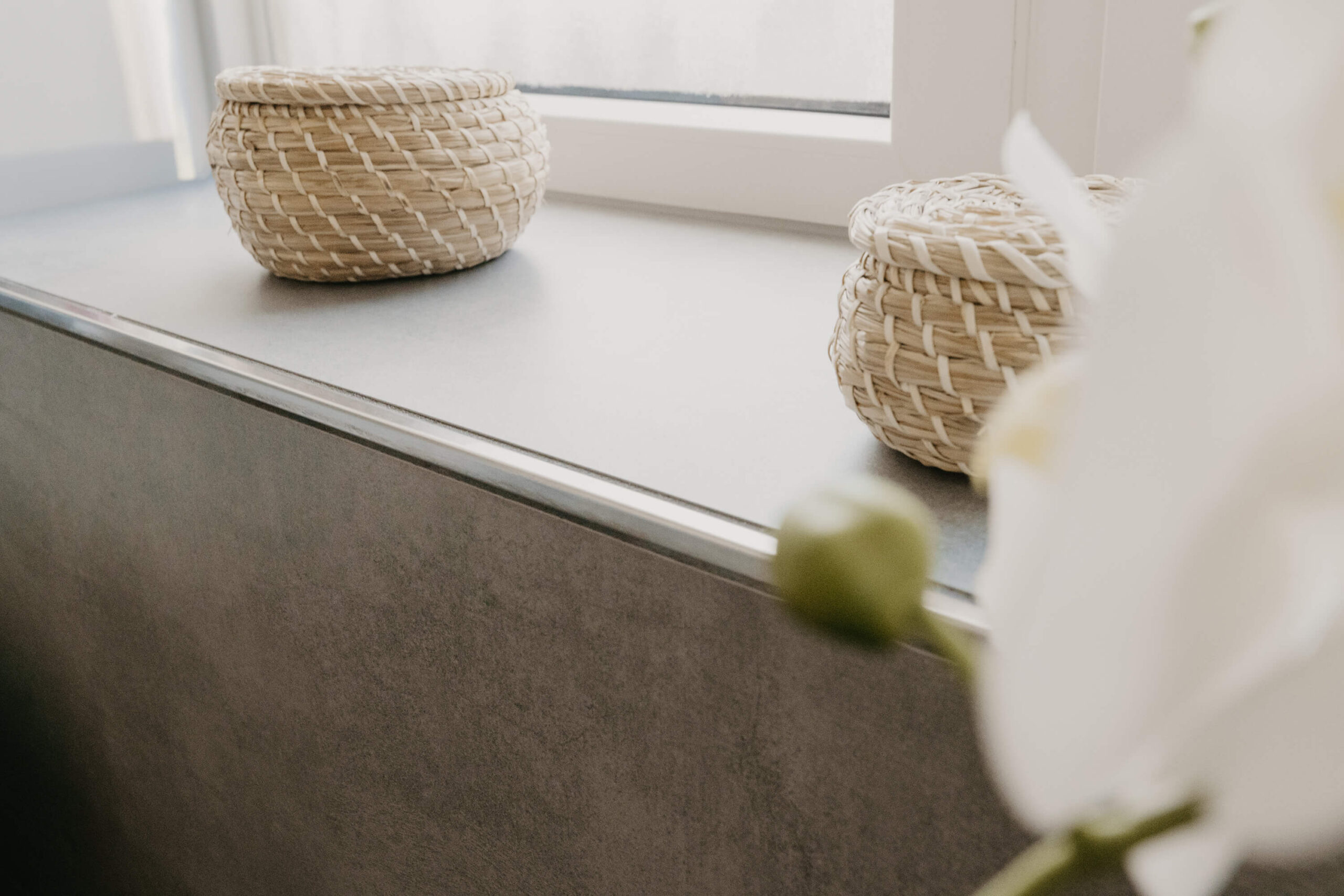 Detailaufnahme einer Fensterbank mit grauer Fliesenoptik, dekoriert mit zwei geflochtenen Körben aus Naturmaterialien.