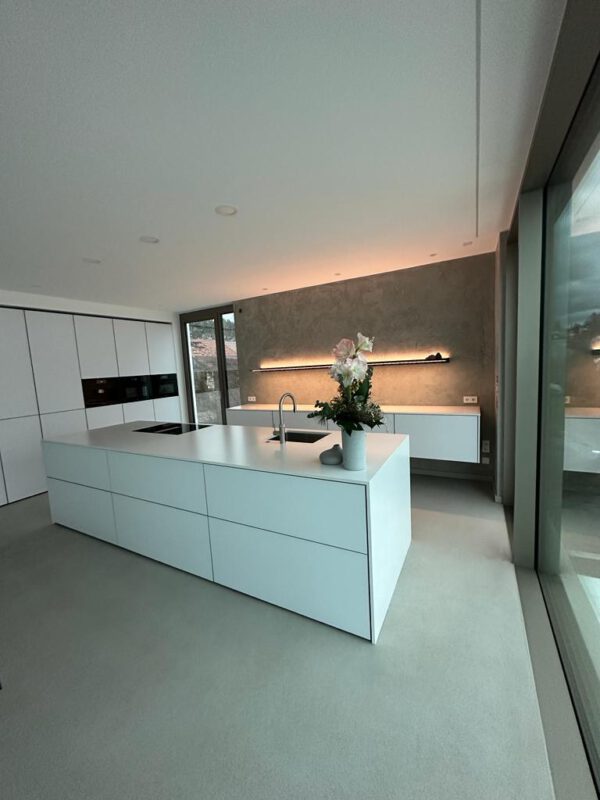 Moderne Küche mit weißer Kücheninsel, minimalistischem Design, Betonoptik-Wand mit indirekter Beleuchtung und großen Fenstern, die viel Tageslicht hereinlassen.