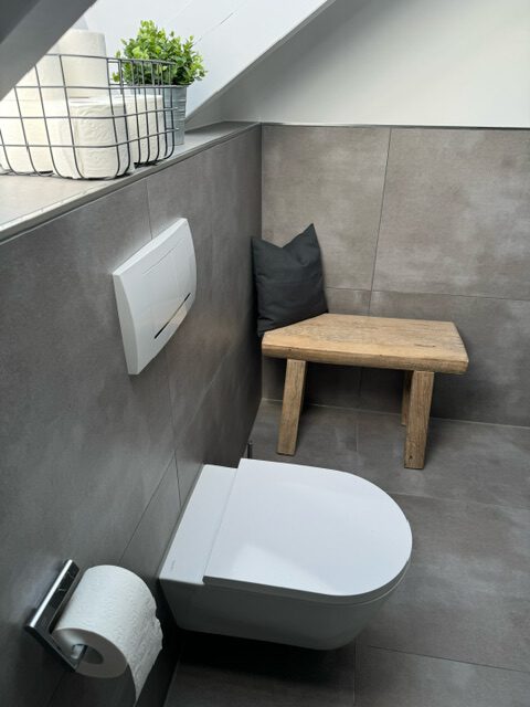 Modernes Gäste-WC mit wandhängendem WC, grauen Fliesen, einer Holzbank mit Kissen und einem Drahtkorb mit Toilettenpapier und einer Pflanze auf der Fensterbank.